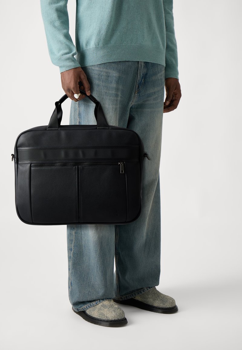 Serviette en cuir noir avec poignées doubles, dotée d'une poche zippée à l'avant et d'une finition texturée. Portée par une personne portant un pull bleu clair et un jean.