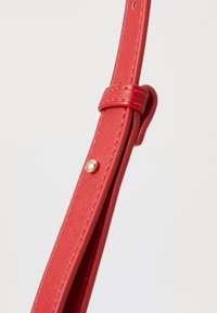 Rode leren band, gladde textuur, verstelbare lengte, verzekerd met een gouden drukknoop, ontworpen voor functioneel gebruik in een tas.