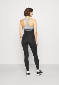 Sutiã desportivo cinzento com faixa sob o logótipo, combinado com leggings pretas de cintura alta. A modelo usa sapatilhas de desporto negras com detalhes brancos.