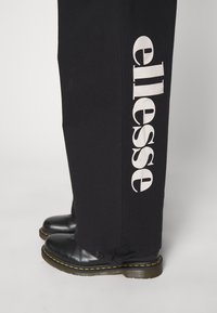Pantalon noir avec un grand logo blanc "ellesse" sur le côté, associé à des bottes noires avec des coutures jaunes et une semelle texturée.