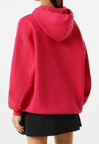 Růžová oversized mikina s kapucí, žebrovanými manžetami a uvolněným střihem, spárovaná s černou sukní s pruhy. Hladká textura a ležérní design.