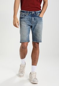 Man som bär urtvättade denimshorts, vita crewstrumpor och beige sneakers, med ena handen i fickan och en vinröd skjorta delvis synlig.