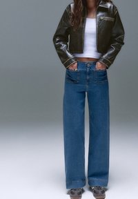 Femme portant une veste courte en cuir noir, un crop top blanc, un jean bleu à jambes larges avec poches avant et des baskets grises, debout les mains dans les poches.