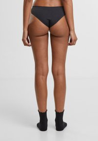 Svart bikini-truser med høy skjæring, glatte stoff og minimal dekning, kombinert med ribbestrikkede svarte ankelstrømper med bølget kant.