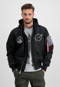 Giacca bomber nera con cappuccio, caratterizzata da toppe e cuciture dettagliate. Chiusura frontale con zip e polsini a coste, abbinata a una maglietta grigia sotto.