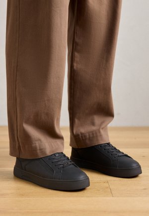 Zwarte lage sneakers van gestept materiaal, rubberen zool en ronde neus. Gecombineerd met losse bruine broeken op een houten vloer.