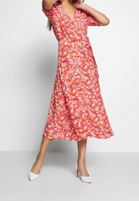 Robe portefeuille rouge à fleurs avec des manches courtes, décolleté en v et taille nouée. Tissu lisse, mi-long, présentant un motif floral blanc et rose éparpillé.