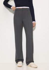 Grijze, op maat gemaakte broek met rechte pijpen, platte voorkant, zijzakken en een vouw langs het been, gecombineerd met lichtgekleurde sneakers.