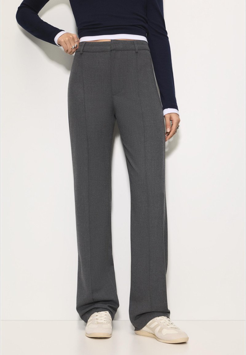 Grijze, op maat gemaakte broek met rechte pijpen, platte voorkant, zijzakken en een vouw langs het been, gecombineerd met lichtgekleurde sneakers.