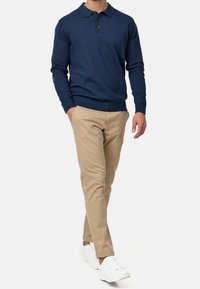 Hombre caminando con una camiseta de polo de manga larga azul marino, pantalones beige, zapatillas blancas, con una mano en el bolsillo frente a un fondo liso.