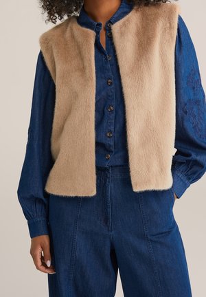 Beige zacht gestructureerd mouwloos vest gedragen over een donkerblauw overhemd met knopen en bijpassende broek met hoge taille.
