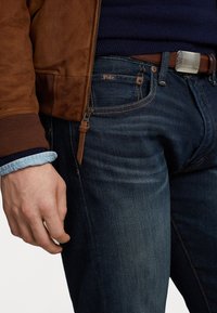 Muž v tmavě modrých džínech s etiketou Polo, hnědým koženým opaskem, námořnickým svetr a hnědou semišovou bundou, s rukou položenou u boku.