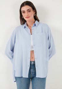 Camicia blu chiaro con bottoni, tessuto strutturato, vestibilità rilassata, maniche lunghe, tasca frontale, indossata sopra un crop top bianco, abbinata a jeans blu.