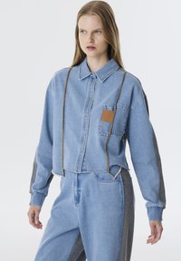 Lichtblauwe denim cropped jas met een knoopsluiting aan de voorkant, een borstzak met een tan lederen patch en grijze stoffen accenten op de mouwen.