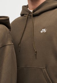 Κοντινό πλάνο ενός ατόμου που φοράει μια φούτερ Nike SB σε ελιά πράσινο χρώμα με κορδόνια και μπροστινή τσέπη.