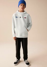 Un sweat à capuche gris clair avec des détails de logo, une poche avant et des poignets côtelés, associé à un pantalon de survêtement noir et des baskets noir et blanc. Un bonnet en tricot bleu est porté.
