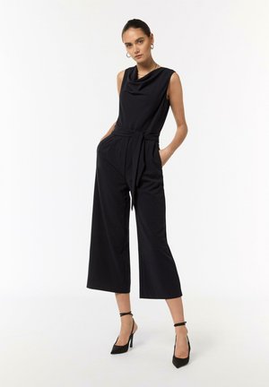 Black sleeveless jumpsuit with draped neckline, wide-leg trousers, and belted waist. Model wearing black high-heeled pumps. Smooth fabric texture.