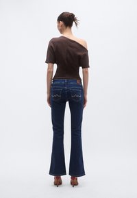 Bruin off-shoulder top gecombineerd met donkerblauwe uitlopende spijkerbroeken. Opvallende details zijn de gebogen achterzakken met gele stiksels en sandalen met hak.