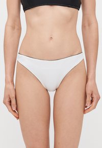 Beli donji dio bikini kostima, niskog struka, s glatkom teksturom, minimalnom pokrivenošću i kontrastnim crnim rubom uz pojas.