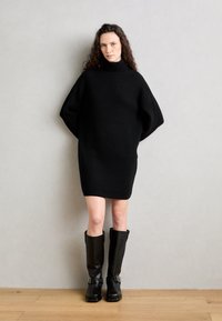 Čierne pletené šaty s vysokým golierom a nadrozmernými rukávmi, doplnené o čierne kožené čižmy po kolená so striebornými prackami.