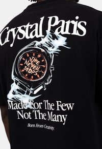Černé bavlněné tričko s grafikou hodinek s oranžovými akcenty. Text zní "Crystal Paris" a "Čas nikdy nezastavuje," s dalšími prvky značky.