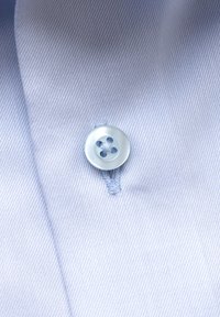 Helehall sinise särgi kanga lähivõte, millel on valge neljarealine nupp õmmeldud nupuaugust kõrgemale.