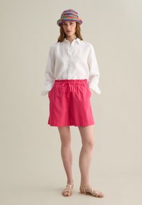Camicia bianca di lino, pantaloni corti rosa con vita elastica, cappello a secchiello colorato in tessuto intrecciato, sandali dorati con cinturini, sfondo neutro.