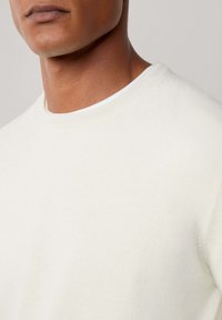 Krupan plan osobe koja nosi jednostavnu bijelu majicu s rundastim izrezom, s vidljivim vratom i donjim dijelom lica, protiv neutralne pozadine.