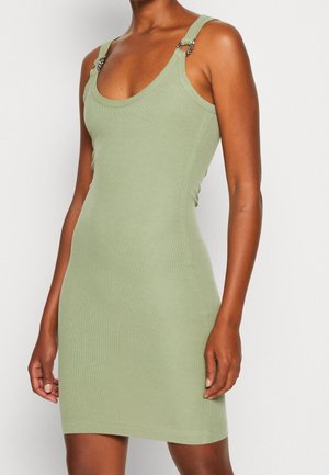 Ärmelloses, hellgrünes, figurbetontes Kleid mit U-Boot-Ausschnitt und dekorativen Metallringen an den Trägern, getragen von einer stehenden Person.