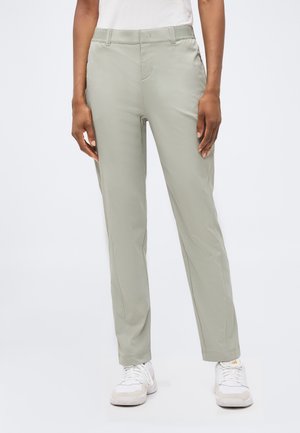 Γυναίκα με ανοιχτό γκρι παντελόνια με ίσια γραμμή και άσπρα αθλητικά παπούτσια, στέκεται μπροστά σε ένα απλό λευκό φόντο.