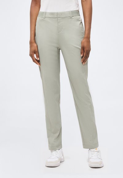 Femme portant un pantalon droit gris clair et des baskets blanches, debout devant un fond blanc uni.