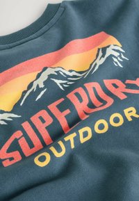 Tessuto blu con una grafica di montagna nei toni del bianco e arancione, con il testo "SUPERDRY OUTDOOR" in lettere grasse rosse e gialle.