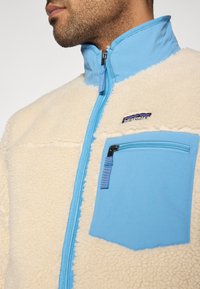Φλις μπουφάν σε κρεμ και ανοιχτό μπλε, με ψηλό γιακά, εμπρόσθιο φερμουάρ και τσέπη στο στήθος με φερμουάρ και λογότυπο.