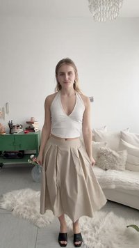 Junge Frau in einem beigefarbenen, plissierten Rock, weißem ärmellosem Oberteil und schwarzen Sandalen, die in einem hellen Wohnzimmer mit grünem Schrank und weißem Sofa steht.