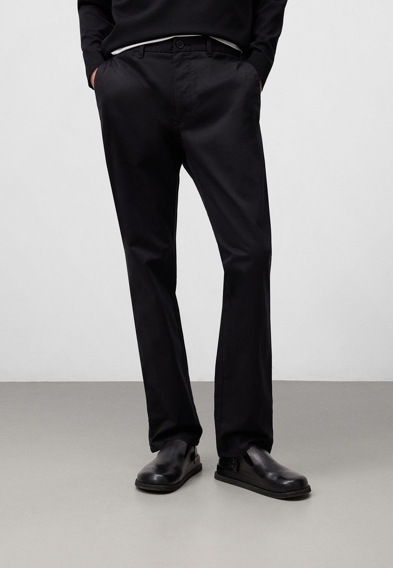 Pantaloni di cotone neri con un design dritto e slanciato, caratterizzati da tasche laterali, passanti per la cintura e una finitura liscia. Abbinati a scarpe nere.