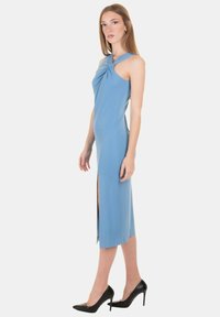 Abito azzurro chiaro, senza maniche, con un design frontale torcigliato, spacco fino al ginocchio e texture liscia; abbinato a tacchi alti neri.
