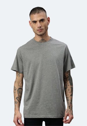 Mann mit kurzen Haaren und Tätowierungen an Armen und Hals, der ein einfaches graues T-Shirt mit kurzen Ärmeln trägt und vor einem hellen Hintergrund nach vorne schaut.