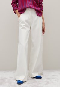 Persona che indossa pantaloni bianchi a gamba larga, una camicia a strisce rosse e blu infilata dentro, e scarpe blu, in piedi contro un muro beige.