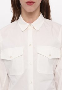 Femme portant une chemise blanche boutonnée avec deux poches à rabat à l'avant et un col pointu, visible des épaules aux lèvres.