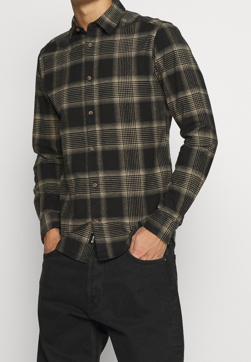 Homme portant une chemise à manches longues boutonnée avec un motif à carreaux noir et beige et un pantalon noir, mains dans les poches, sur un fond uni.