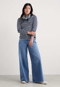 Kobieta ubrana w granatową i białą w paski bluzkę z długim rękawem, niebieskie szerokie dżinsy, białe trampki i wzorzystą chustę zawiązaną na szyi.