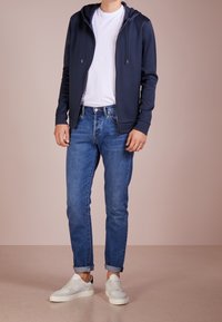 Marinefarbener Reißverschluss-Hoodie über einem weißen T-Shirt, kombiniert mit hochgekrempelten blauen Jeans und weißen Sneakers. Glatter Stoff, lässiger Schnitt und minimalistisches Design.