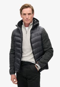 Gilet imbottito nero con maniche in maglia grigia testurizzata, dotato di cappuccio e chiusura con zip. Indossato su un maglione grigio chiaro con zip a quarto.