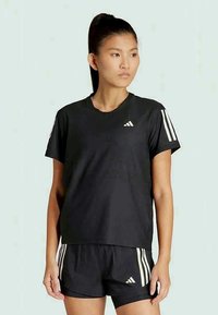 Atletska ženska v črni Adidas športni opremi, ki vključuje majico kratkih rokavov in kratke hlače z belimi črtami, stoji pred enobarvno ozadjem.