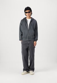 Graue Zip-Hoodie mit Fronttaschen, kombiniert mit lockeren, dunkelgrauen Hosen und weißen Sneakers. Das Model trägt Sonnenbrillen und ein weißes T-Shirt.