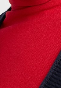 Maglione a collo alto rosso con una texture liscia, parzialmente coperto da un gilet di maglia scuro. Focus sul colore vibrante e sui dettagli di cucitura curati.