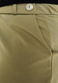 Khaki byxor med ett slätt, texturerat tyg. En rund, krämfärgad knapp säkrar midjan, med snedställda framfickor synliga.