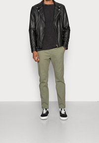 Giacca in pelle nera da motocicletta sopra un maglione lavorato a maglia scuro, abbinata a pantaloni khaki e sneakers nere con suole bianche.