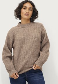 Maglione lavorato a maglia color marrone chiaro con collo alto e spalle scese, caratterizzato da un motivo testurizzato e polsini a costine. Abbinato a jeans scuri.