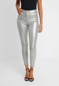 Jeans skinny metalizados plateados de talle alto con una textura suave, con cierre de botón en el frente y cinco bolsillos, combinados con sandalias negras.